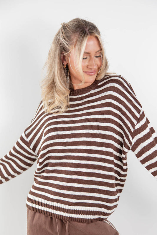 A woman wearing a brown and off-white horizontally striped knit sweater, shown from the waist up against a plain light gray studio background.
