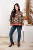 A woman modeling a long-sleeve mock neck sweater with a dark olive green base and a vibrant pink, red, and white floral pattern, paired with dark blue jeans and brown heeled boots.