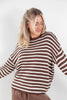 A woman wearing a brown and off-white horizontally striped knit sweater, shown from the waist up against a plain light gray studio background.