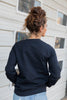 A woman with her hair in a messy bun from behind wearing a black crewneck sweatshirt paired with light blue jeans, standing in front of a white house exterior.