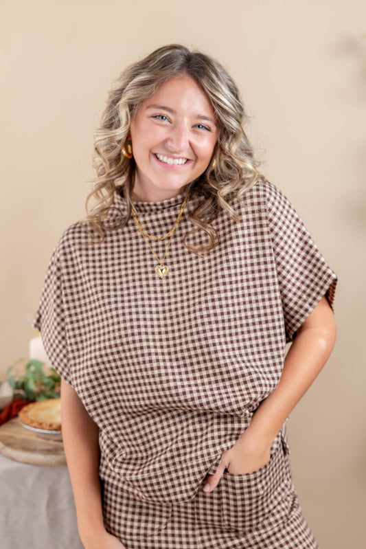 A smiling woman with blonde-highlighted curly hair wearing a brown and beige gingham short-sleeve top and matching skirt, standing against a warm beige background.