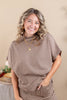 A smiling woman with blonde-highlighted curly hair wearing a brown and beige gingham short-sleeve top and matching skirt, standing against a warm beige background.