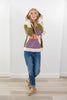 A woman modeling a color-blocked knit hoodie with olive green, purple, cream, and orange panels, paired with blue jeans and tan loafers, standing in a bright room with white walls and light wood flooring.