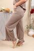 A woman wearing brown and beige gingham wide-leg pants with ruffled hems, paired with tan slip-on shoes, standing in a styled indoor room with a table and decorative plants.