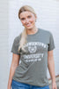 A woman with stands in front of a white brick wall wearing a olive green-colored graphic t-shirt with the words 'Halloweentown University, Class of '98' printed in white text. She is also wearing blue jeans.