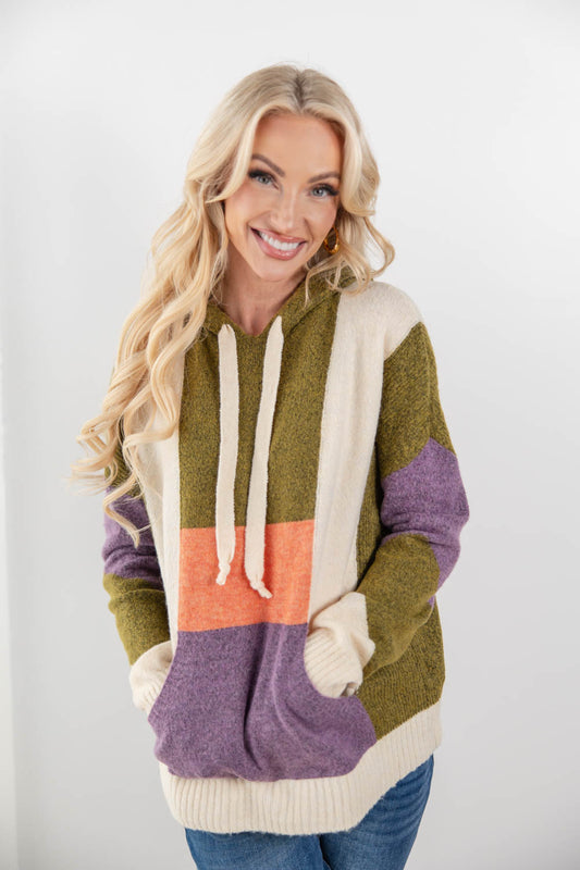 A smiling blonde woman wearing a chunky color-block knit hoodie with olive green, cream, purple, and orange panels, paired with blue jeans, against a plain light gray studio background.