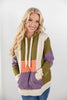 A smiling blonde woman wearing a chunky color-block knit hoodie with olive green, cream, purple, and orange panels, paired with blue jeans, against a plain light gray studio background.