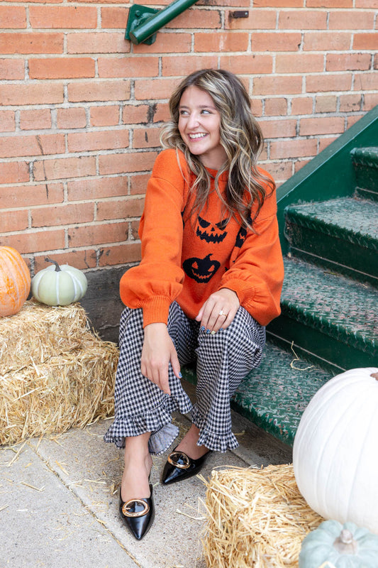 A woman sits on outdoor steps next to a brick wall, wearing a bright orange Halloween sweatshirt with a black jack-o'-lantern graphic, black and white gingham pants, and black pointed flats.