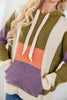 A close-up of a woman wearing a chunky knit color-block hoodie with olive green, cream, purple, and orange panels, featuring cream drawstrings and a front kangaroo pocket.