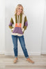 A woman modeling a color-blocked knit hoodie with olive green, cream, orange, and purple panels, paired with blue jeans and tan loafers, standing against a white wall.
