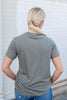 A rear view of a woman with standing in front of a white wearing a olive green-colored graphic t-shirt with blue jeans.