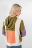 A woman shown from the back wearing a color-block knit hoodie with olive green, cream, peach, and purple panels, paired with blue jeans against a light gray studio background.