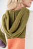 A close-up of a person wearing a chunky knit colorblock hoodie. The hoodie features a large olive green hood, a white body, and a peach-orange colorblock panel at the bottom.