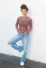 A woman posing against a white wall wearing a long-sleeve mock neck top with a black and orange checkerboard pattern, light blue distressed jeans, and white sneakers.