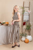 A woman modeling a sleeveless midi dress with vertical brown and beige stripes, standing in a styled indoor autumn setting with pumpkins, a table, and a ladder backdrop.