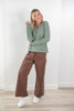 A woman modeling a green, brown and white striped long-sleeve top with brown wide-leg pants featuring white side stripes, paired with white sneakers, standing in a minimalist room with a white wall and light wood floor.