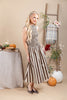 A woman modeling a sleeveless midi dress with vertical brown and cream stripes, standing in a styled studio setting with pumpkins, a table, and a ladder backdrop.