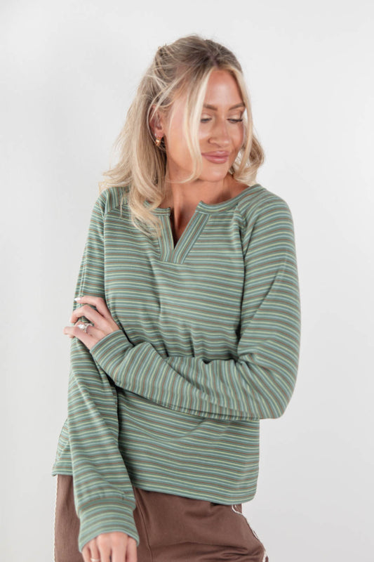 A woman wearing a sage green, brown and white striped long-sleeve brushed henley shirt, paired with brown pants, standing against a plain light gray background.