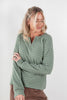 A woman wearing a sage green, brown and white striped long-sleeve brushed henley shirt, paired with brown pants, standing against a plain light gray background.