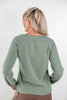 A woman shown from the back wearing a sage green long-sleeve raglan top with thin horizontal tan and white stripes, paired with brown pants, against a light gray studio background.