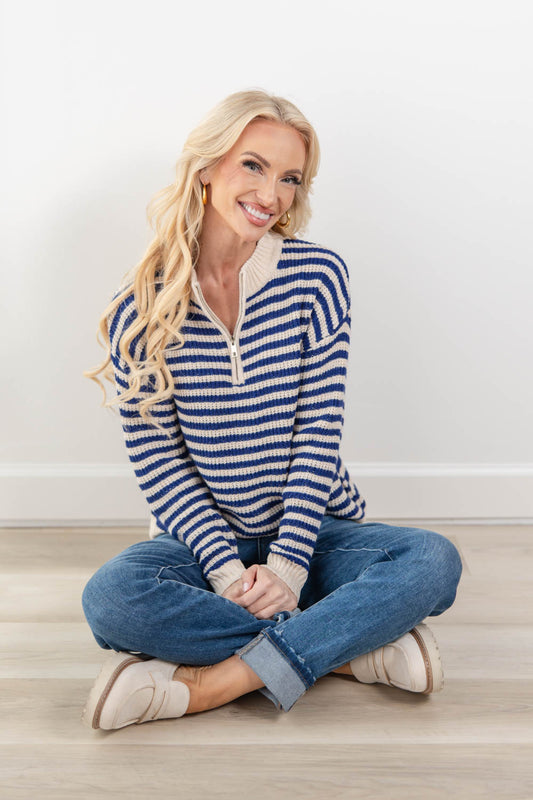 A smiling blonde woman sits cross-legged on a light wood floor against a white wall, wearing a blue and cream striped quarter-zip knit sweater, blue jeans, and beige slip-on shoes.