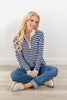 A smiling blonde woman sits cross-legged on a light wood floor against a white wall, wearing a blue and cream striped quarter-zip knit sweater, blue jeans, and beige slip-on shoes.