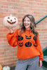 A woman wearing a bright orange crew neck sweater with a black jack-o'-lantern graphics, paired with black and white gingham ruffled pants as she holds a jack-o-lantern.