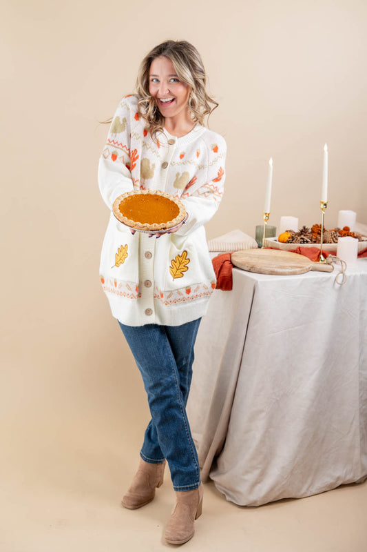 A woman stands in a studio setting holding a pumpkin pie, wearing a white button-front cardigan with orange and yellow autumn leaf patterns, blue jeans, and tan ankle boots. A draped table with candles and fall decor is visible to her right against a warm beige backdrop.