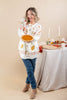 A woman stands in a studio setting holding a pumpkin pie, wearing a white button-front cardigan with orange and yellow autumn leaf patterns, blue jeans, and tan ankle boots. A draped table with candles and fall decor is visible to her right against a warm beige backdrop.