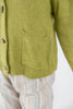 Close-up of a textured olive green knit cardigan with brown wood toggle buttons and a front patch pocket, worn over light beige and blue striped trousers.