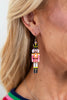 A close-up of a woman's ear wearing a dangling nutcracker soldier earring. The earring features a colorful painted nutcracker figure with a gold top hat and silver-toned hoop earring.