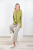 A woman sitting on a white stool wearing a sage green cardigan and beige and blue toned striped wide-leg pants, photographed in a bright indoor room with light wood flooring and white walls.