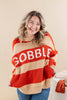 A woman smiling while wearing a chunky knit sweater with bold horizontal stripes in beige and rust red, featuring the word 'GOBBLE' in large white letters across the chest. She is also wearing blue jeans, posed against a solid beige studio background.