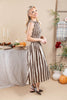 A woman modeling a sleeveless midi dress with vertical brown and cream stripes, standing in a styled indoor setting with autumn decorations.