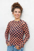 A woman wearing a long-sleeve checkered top with a black and orange checkerboard pattern and small jack-o-lantern details, paired with light blue jeans against a white textured wall.