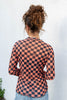 A woman viewed from behind wearing a long-sleeve checkered top with a black and peach-orange checkerboard pattern and small jack-o-lantern details, paired with light blue jeans against a white textured wall.