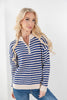 A woman with blonde hair wearing a blue and cream striped quarter-zip sweater and blue jeans, standing against a plain white wall.