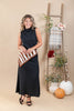 A woman stands in a studio wearing a sleeveless black midi dress and holding a brown and white striped scarf. The background features a beige backdrop, a wooden ladder decorated with a plaid throw blanket, flowers, and potted pumpkins.