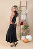 A woman modeling a sleeveless black maxi dress with a brown and white striped waist tie, standing in a studio decorated with a wooden ladder, pumpkins, and vases.