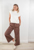 A woman modeling a white ribbed knit mock neck sleeveless top and brown wide-leg drawstring pants with white side stripes, paired with beige slip-on shoes, standing against a white wall.