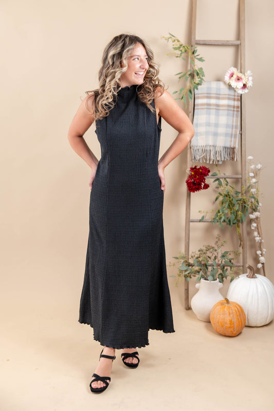 A woman modeling a sleeveless, mock neck, floor-length black dress with a textured waffle-like fabric, standing in a warm beige studio decorated with a wooden ladder, pumpkins, and potted plants.