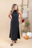 A woman modeling a sleeveless, mock neck, floor-length black dress with a textured waffle-like fabric, standing in a warm beige studio decorated with a wooden ladder, pumpkins, and potted plants.