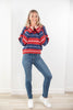A woman modeling a red, navy, and white striped knit sweater, paired with blue jeans and white sneakers, standing against a white wall.
