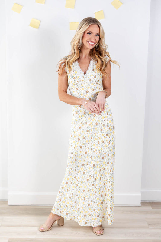 A woman modeling a sleeveless white maxi dress with a yellow and brown floral print, standing in front of a white wall and light wood floor.