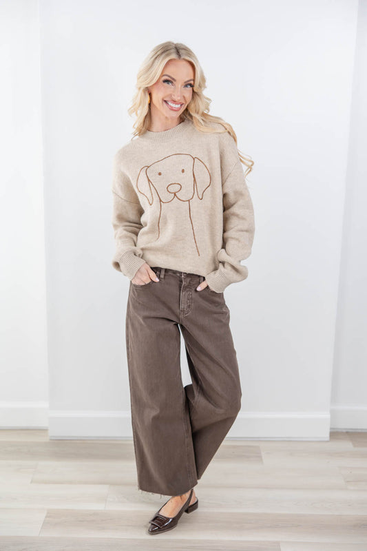 A woman modeling a beige crewneck sweater with a brown line art dog graphic, paired with brown wide-leg pants and dark brown flats, standing in a bright room with white walls and light wood floors.