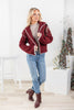 A woman modeling a burgundy knit cardigan with white trim, styled with a beige turtleneck, blue jeans, and burgundy ankle boots.
