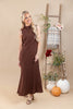 A woman modeling a sleeveless, high-neck, floor-length brown dress with a textured waffle-like fabric, standing in a studio decorated with a wooden ladder, pumpkins, and potted plants.