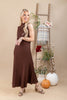 A woman models a sleeveless, high-neck, floor-length brown dress with a textured knit pattern. She is standing in a studio setting with a beige background, next to a wooden ladder decorated with a plaid throw blanket, flowers, and pumpkins.