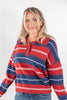 A woman wearing a red, navy blue, and white striped knit polo sweater with blue jeans, photographed against a plain light gray studio background.