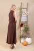 A woman modeling a sleeveless, high-neck brown maxi dress with a subtle textured pattern, standing in a studio decorated with a wooden ladder, pumpkins, and vases.